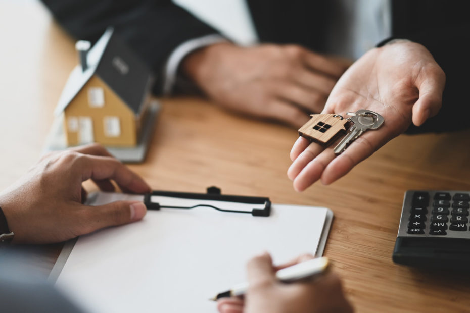 Cropped shot of Real estate agent giving a property key to his customer while signing an agreement on clipboard at the modern wooden table. Realtor/Broker/Seller/Dealer concept.